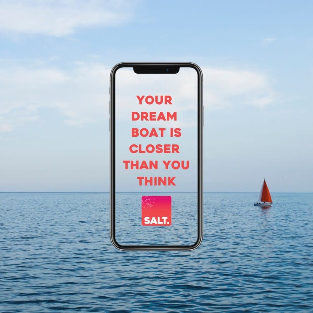 A phone showing the SALT app slogan “Your dream boat is closer than you think”, against a calm sea with a sailing yacht on the horizon